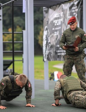 Dwie osoby w mundurach wojskowych podczas ćwiczenia - pompki. Nad nimi stoi inna osoba w mundurze wojskowym i czerwonym berecie.
