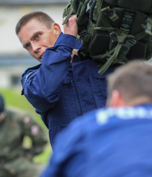 Osoba w mundurze policyjnym niesie na ramieniu ciężki duży plecak wojskowy.