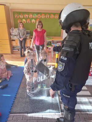 Policjant ubrany w kamizelkę przeciwuderzeniową, ochraniacze nóg i kask prezentuje dzieciom wyposażenie typowe dla funkcjonariuszy oddziałów prewencji.