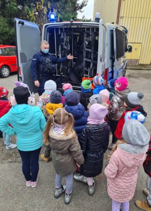 Policjant prezentuje zaciekawionym przedszkolakom tylną zabudowę radiowozu typu furgon.
