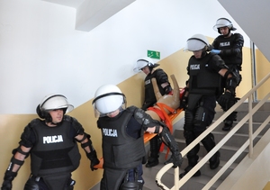 Policjanci w sprzęcie PZ znoszą manekina na desce po schodach koszarowca.