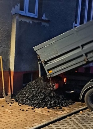 Wywrotka wysypująca węgiel przy ścianie budynku.