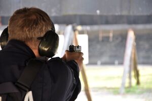 Policjant oddaje strzał. Ujęcie od tyłu. Zbliżenie na broń. W tle rozmazana tarcza i kulochwyt.