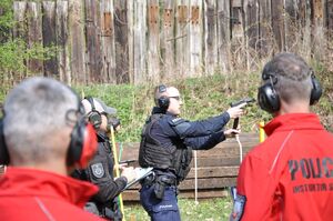 Policjant wymienia magazynek podczas konkurencji strzelania do gongu. Na pierwszym planie rozmazani sędziowie. Ujęcie z boku.