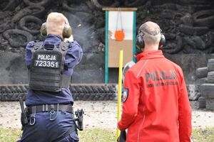 Policjant i sędzia podczas konkurencji strzelania do gongu. Widoczna smuga dymu i łuska w powietrzu. Ujęcie od tyłu.