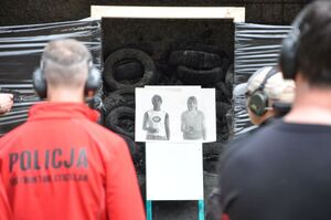 Tarcza przedstawiająca kobietę z bronią obok tarczy neutralnej kobiety z latarką - w tle wyostrzone. Na pierwszym planie rozmazani sędziowie zawodów.