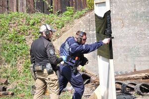 Policjant oddaje strzał prawą ręką zza przesłony. Za nim sędzia zawodów. Ujęcie z boku.
