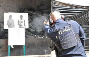 Policjant oddaje strzał lewą ręką zza przesłony do tarczy przedstawiającej kobietę z bronią obok tarczy neutralnej. Widoczna chmura dymu i łuska w powietrzu. Ujęcie od tyłu.