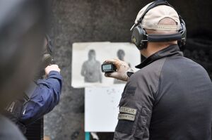 Na pierwszym planie sędzia zawodów ze specjalistycznym timerem strzeleckim. W tle tarcze strzeleckie.