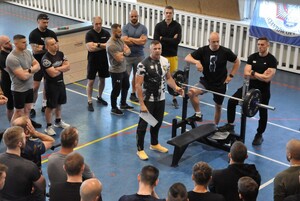 Jeden z sędziów prezentuje stanowisko do pierwszej konkurencji wyciskania sztangi. Obok pozostali sędziowie i zawodnicy.