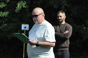 Główny sędzia zawodów oficjalnie rozpoczyna drugi dzień zmagań. W tle jeden z sędziów i tarcza strzelnicza.