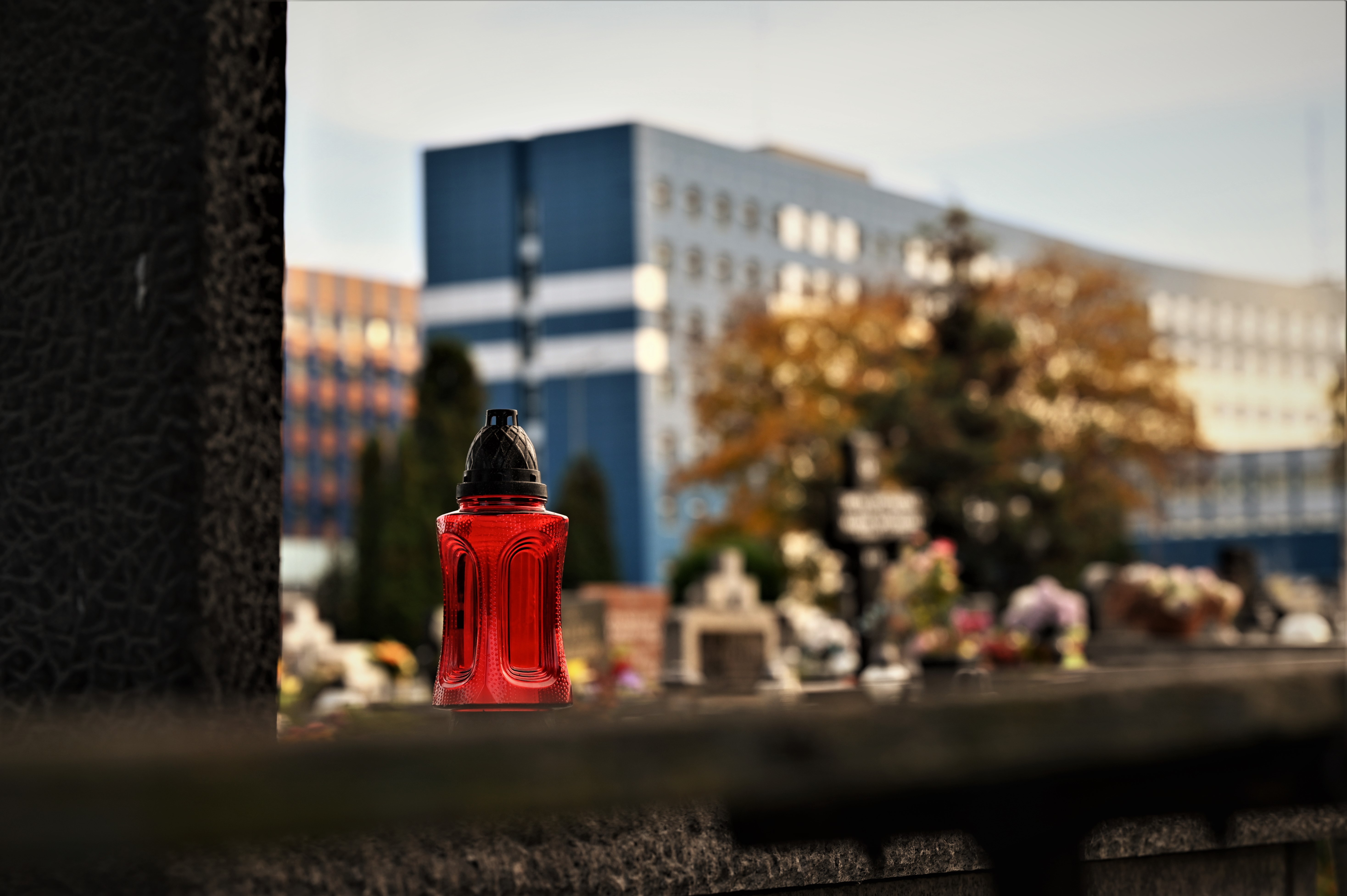 zdjęcie przedstawia zbliżenie na znicz stojący na grobie, w tle budynek Komendy Wojewódzkiej Policji w Katowicach